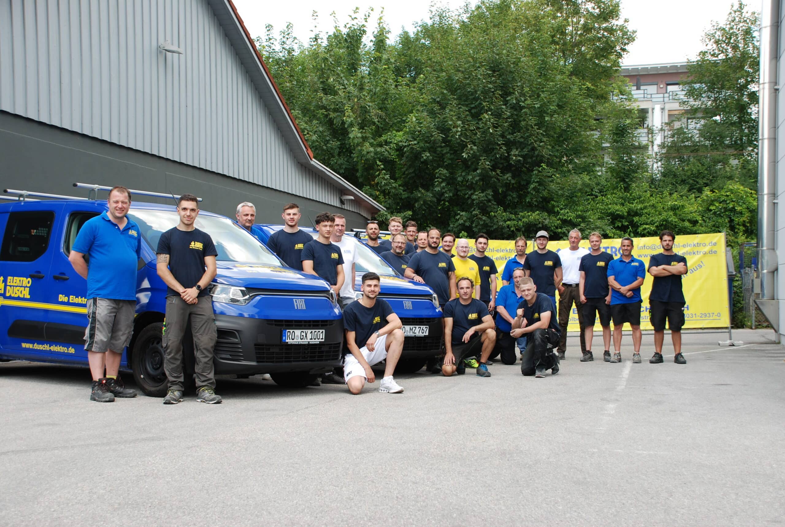 Teamfoto Das Team von Elektro Duschl steht zu 25 Personen vor drei ihrer blauen Firmenwägen
